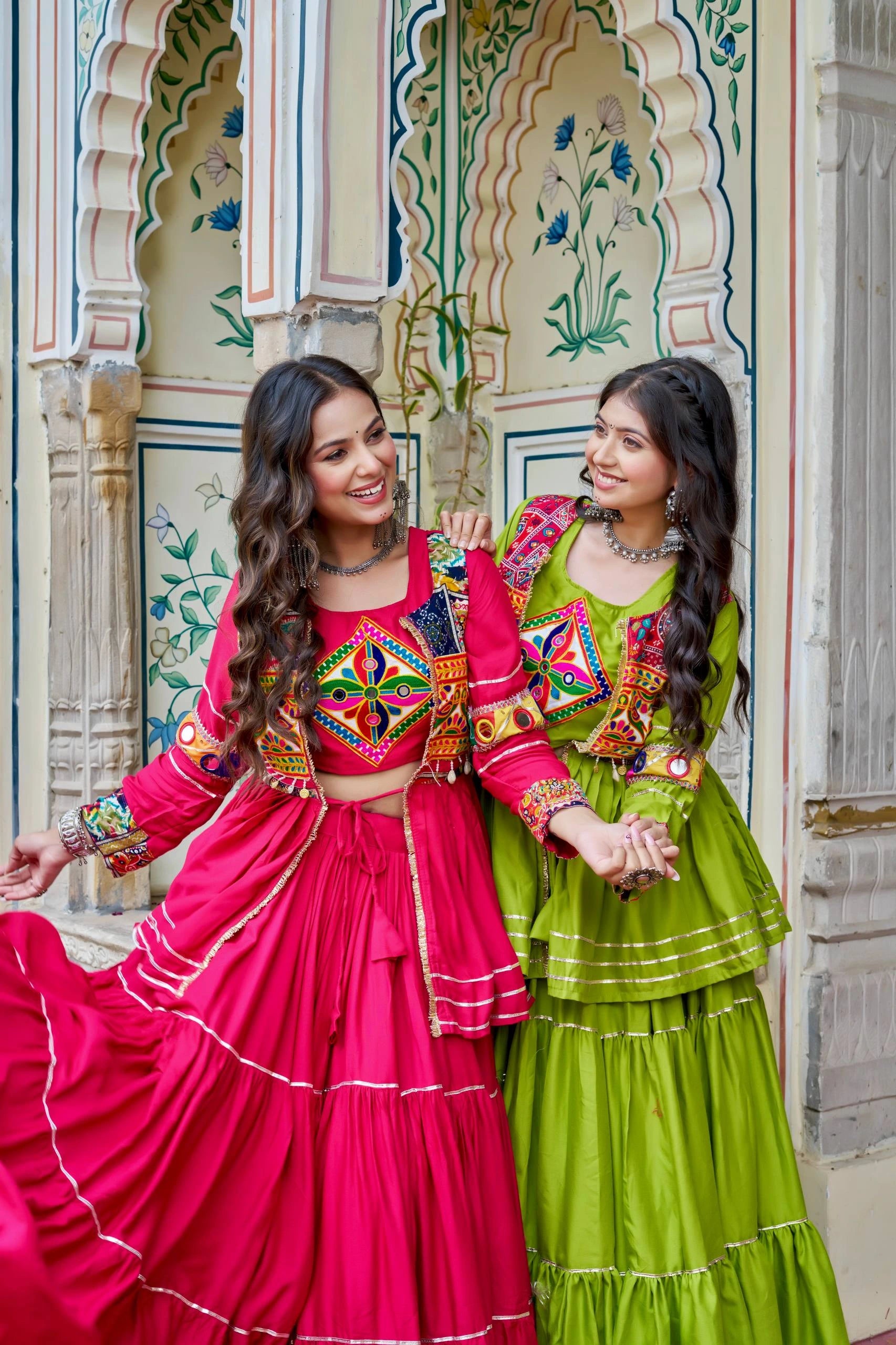 Two women in colorful traditional outfits standing in front of an ornate architectural background.